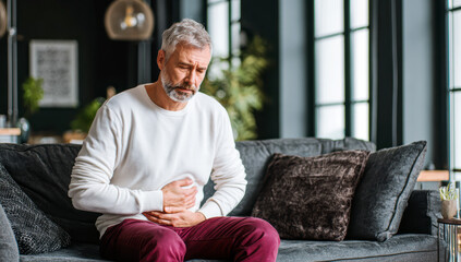 Fototapeta premium Mature man holding his stomach, experiencing intense pain and discomfort due to indigestion or illness at home