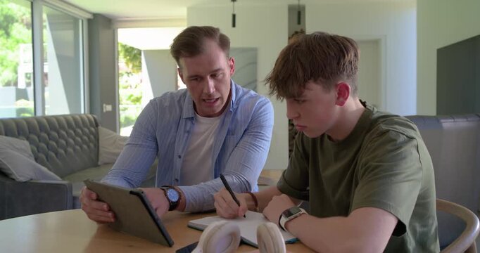 Father and adolescent son at wooden table, father angling tablet, son writing notebook for homework