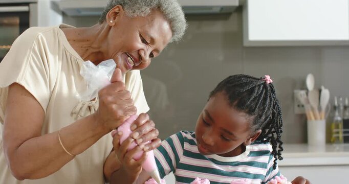 Senior and Youth African women holding piping bag guiding piping pink swirls on cupcakes in kitchen