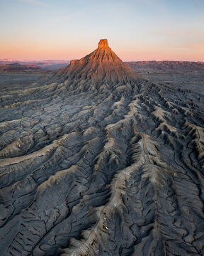 Aerial view of the Factory Butte peak bathed in golden light, its rugged, grey slopes contrasting with the soft peach hues of the sky, Utah, United States.