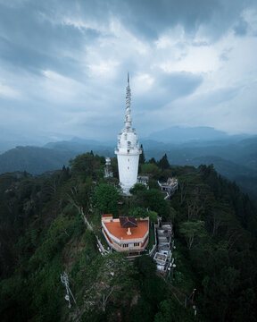 Aerial view of the white Ambuluwawa Tower piercing the sky above verdant hills, its spiral form a stark contrast against the moody blues, Gampola, Central Province, Sri Lanka.
