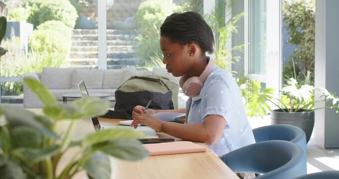 African female youth sitting at table at home, glancing at laptop and writing notes while studying