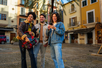 Obraz premium Diverse group of young adult friends enjoying urban travel, smiling and navigating a foreign city street together with a smartphone, finding directions while exploring