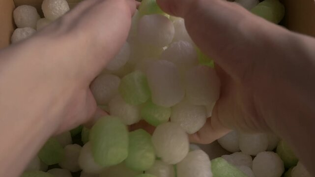 Hands sorting ecofriendly packing peanuts in a cardboard box, showcasing green and white biodegradable materials for sustainable packaging. Ecofriendly packaging