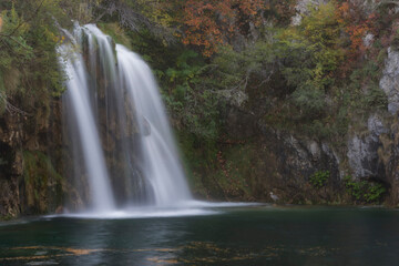 Obraz premium Professional long exposure of a waterfall cascading into a dark pond. Features autumn foliage, rocky textures, and a small heron. High-resolution nature photography for travel and editorial use.
