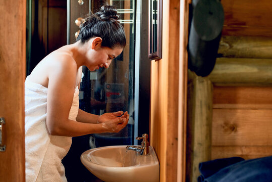 Woman in a towel cleansing her face at a bathroom sink in a rustic wooden cabin. Self care, hygiene and natural beauty routine indoors.
