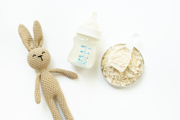 Powdered infant formula in jar lid and scoop with baby milk bottle and knitted bunny on white background. Top view.