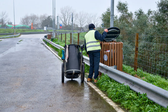 Maintenance worker emptying a wooden trash bin on a rainy roadside.