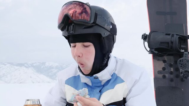 Young skier bites sweet blue glazed donut while sitting on snack break in snowy mountains. Snowboarder eats pastry and drinks hot beverage from thermal mug. Outdoor athlete snack.