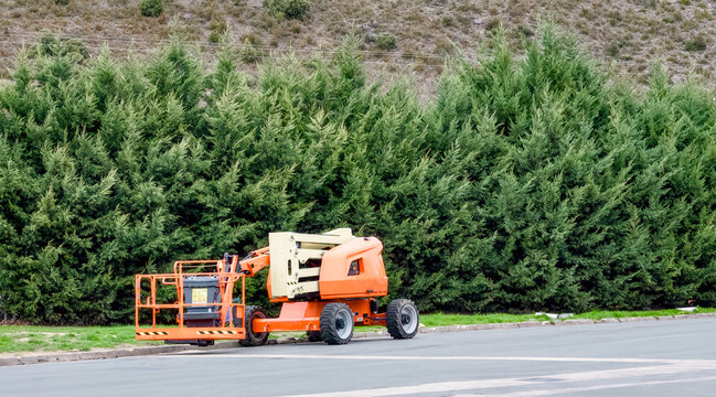 Orange articulated boom lift parked on a roadside against a green hedge.
