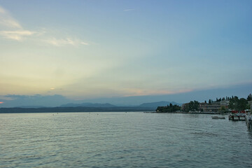 A peaceful evening view of the calm waters of Lake Garda near Sirmione, featuring a soft sunset sky, distant mountains, and coastal buildings.