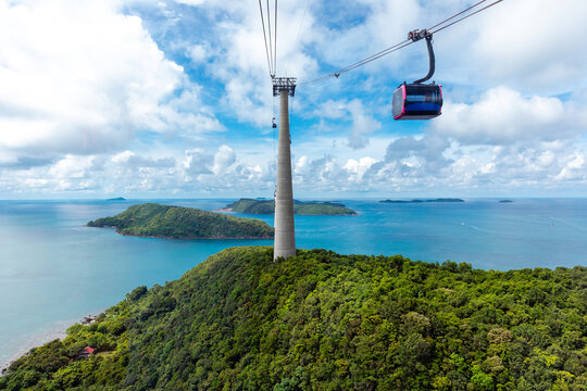 Aerial view of a cable car traveling over green hills and turquoise waters, with distant islands visible under blue sky. The world's longest cable car system, connecting Phu Quoc with Hon Thom island