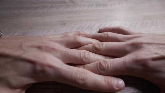 A close-up shot of two hands slowly coming together, interlacing their fingers on a wooden table. Concept of contemplation, anxiety, connection, intimacy, and human relationships.