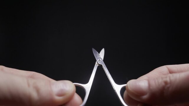A person's hands hold a pair of small, metal nail scissors, repeatedly opening and closing them against a dark background. Concept of personal grooming, hygiene, and self-care.