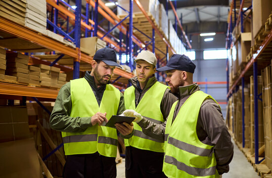 Warehouse logistics inventory workers using tablet. Crew in safety vests review orders and stock by racks aligning loading and delivery. Collaborative warehouse operations and logistics concept.