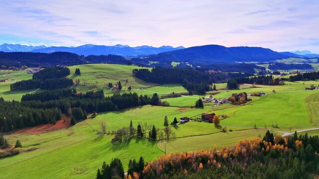 Aerial view of green meadows and forests in Bavaria. Rolling green hills with pine forests, small farm houses and snow capped Alps in distance.