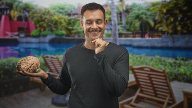 Man holding a model brain in left hand, smiling with hand to chin gesture at building poolside deck; curiosity thinking.