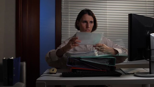 Woman working late in a dim office, hunched over stacks of paperwork and documents, exhausted and stressed by heavy workload, deadlines and long hours of focused effort