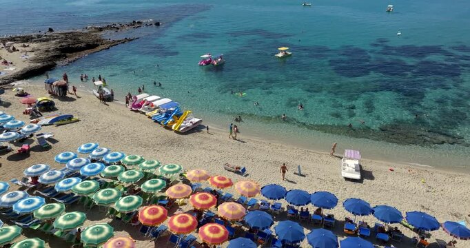 Aerial view of a Mediterranean beach featuring organized rows of blue and striped yellow-green umbrellas. Swimmers enjoy the crystal-clear turquoise water that gently laps against the pale sandy shore