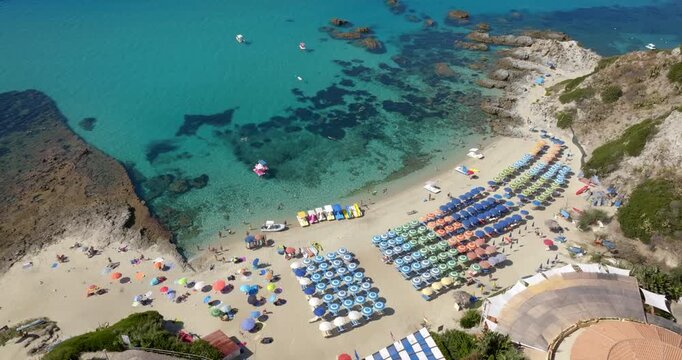 Aerial view of a crowded Mediterranean beach featuring dense rows of colorful sun umbrellas. The crystal-clear turquoise water reveals dark rocky reefs near the sandy shoreline. It's a summer morning.
