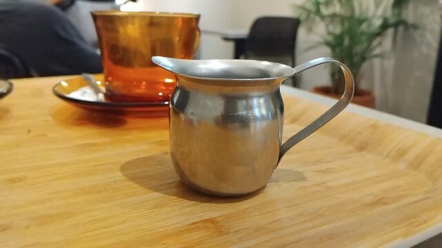 Close-up of a small vintage metal cream pot on a wooden caf&eacute; table in France, with a transparent yellow cup nearby, retro and slightly kitsch atmosphere.
