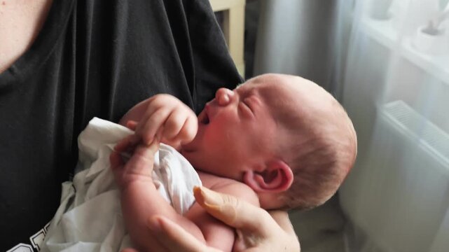 A week-old baby cries in his mother's arms. Soft natural light illuminates the baby's flushed face. A casual, everyday moment of early parenthood. Close-up