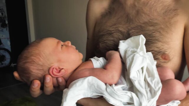 A tender close-up of a father cradling his newborn baby. The infant is wrapped in a white sheet. The intimacy of skin-to-skin contact and paternal affection are evident. Natural light