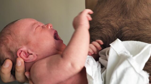 A close-up profile captures the raw emotion of a crying week-old newborn. With flushed skin and an open mouth, the infant is held safely in a parent's arms. Soft focus