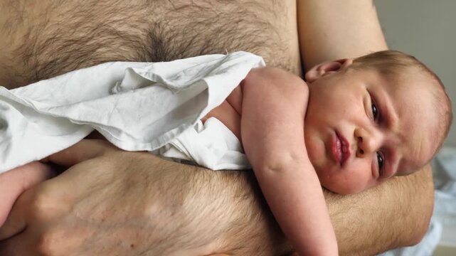 A father holds his newborn infant, wrapped in a white cloth, against his bare chest. Close-up shot with soft lighting captures an intimate moment of love, bonding, and paternal care