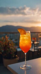 A vibrant cocktail with an orange slice garnish in a tall glass sits on a table. The background features a sunset view over distant mountains and a partially blurred balcony setting