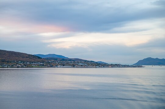 Tranquil fjord waters reflect dramatic cloudy sky in Sortland, Norway. Mountain landscape with coastal town creates peaceful Nordic scenery perfect for travel and nature content.