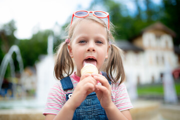 Little girl licking vanilla ice cream cone with red sunglasses on head in park, fountain bokeh background