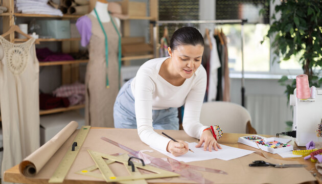 Creative female seamstress designer drawing sketches on paper while sewing dress at workplace in small fashion sewing workshop