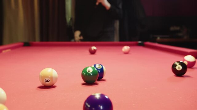 Young caucasian adult male player hits balls by cue. Closeup of striking clustered balls. Youthful player shatters balls on red felt table with lively night club ambiance. Match in a competitive sport