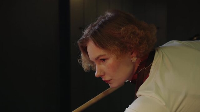 Portrait of focused young caucasian woman with steady grip holds the cue tightly and hits billiard ball. Precise trajectory and calm, solo training session with competitive warmup vibe in hight club