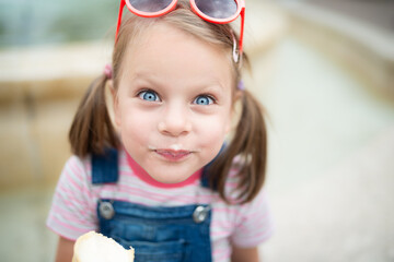 Close-up portrait of cute little girl with blue eyes and red sunglasses on head eating vanilla ice cream cone, milk moustache, soft bokeh background