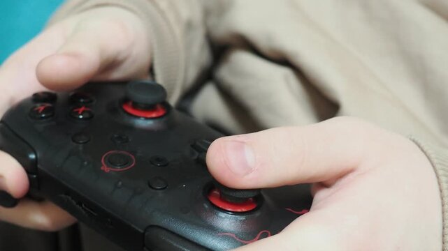 Concentrated boy in big gaming headphones playing video game with wireless controller console joystick, sitting on bed. Concept of gaming, children and technology, screen time, and leisure at holidays