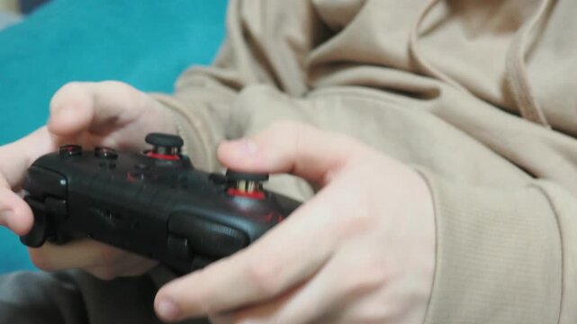 Concentrated boy in big gaming headphones playing video game with wireless controller console joystick, sitting on bed. Concept of gaming, children and technology, screen time, and leisure at holidays