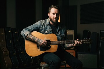 Obraz premium A portrait of a bearded musician with tattoos on his arms, sitting with his acoustic guitar in a music studio.