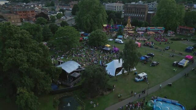 An aerial view shows a lively outdoor fairground event in a park, with carnival rides, food stalls, a stage, and a large crowd enjoying the festivities.