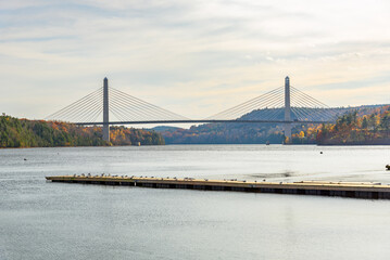 Obraz premium Modern suspension road bridge spanning a large river with forested banks at sunset in autumn. An empty wooden jetty is in foreground.