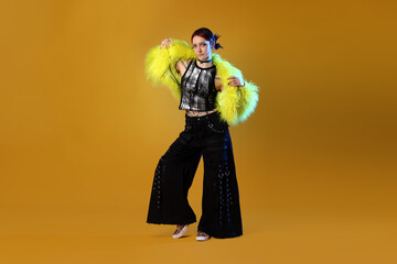 Beautiful young woman with piercings in fur coat and wide pants posing in color light on orange background