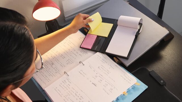 Young woman placing a sticky note on her notebook to highlight an important date, representing organization, planning, reminders, productivity, and daily work routine.