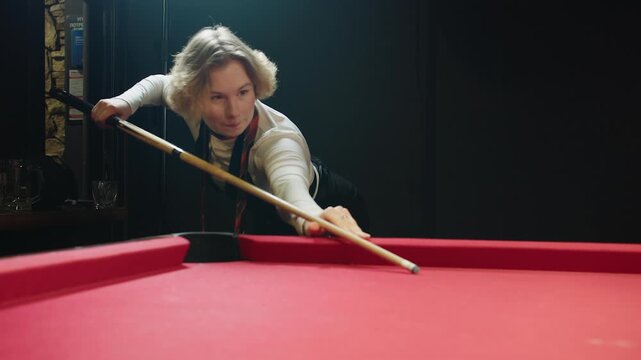 Young adult caucasian woman in casual cloth leaning over red pool table and holds cue, careful aiming for single white ball in dim club lighting and focused practice session showcasing skill.
