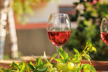 Glass of rose wine with white grapes and vine leaves on wooden table with garden and house in background © svittlana