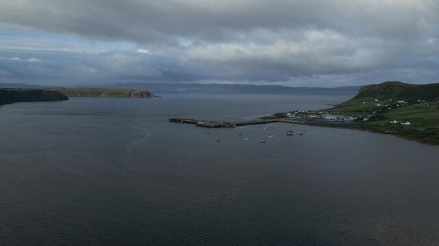 A coastal village with a pier and boats sits beside a calm sea, with dramatic cloudy skies overhead and distant hills.