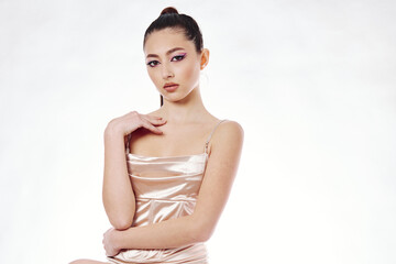 Elegant young woman with slicked back hair in a shiny satin dress poses confidently against a white...