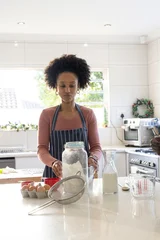 Fotobehang Koken African American woman at kitchen island wearing striped apron preparing with flour jar and eggs  © wavebreak3
