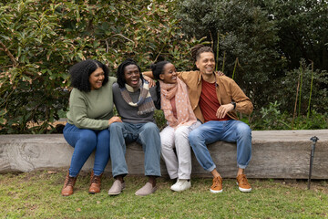Diverse friends sitting closely on wooden log bench in park garden, wearing scarves, smiling