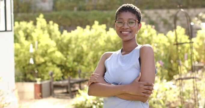 Adult female standing arms crossed in sunny garden, wearing specs, tank top sunlight widening smile